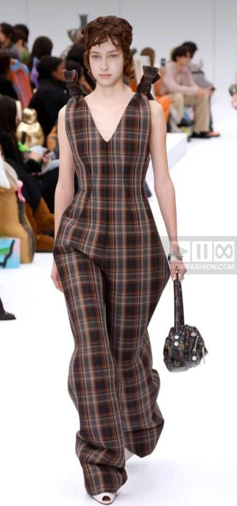 “Model showcasing a brown plaid structured jumpsuit with a deep V-neckline and matching handbag on the runway.”