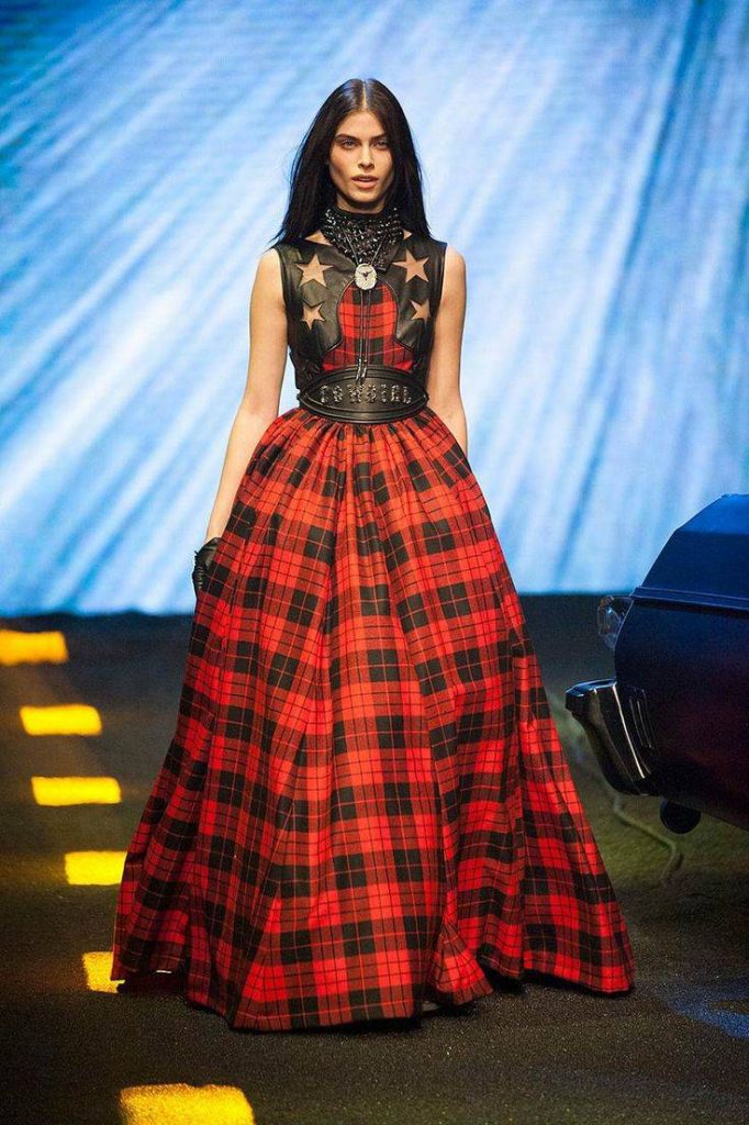 Plaid Runway Jumpsuit: “Model showcasing a brown plaid structured jumpsuit with a deep V-neckline and matching handbag on the runway.”