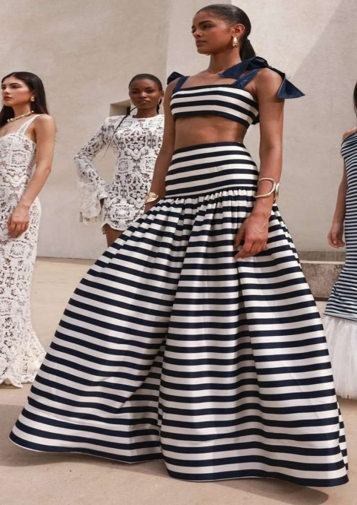 : “Model in a black and white striped maxi two-piece dress with bow shoulder straps, walking the runway alongside lace-detailed dresses.”