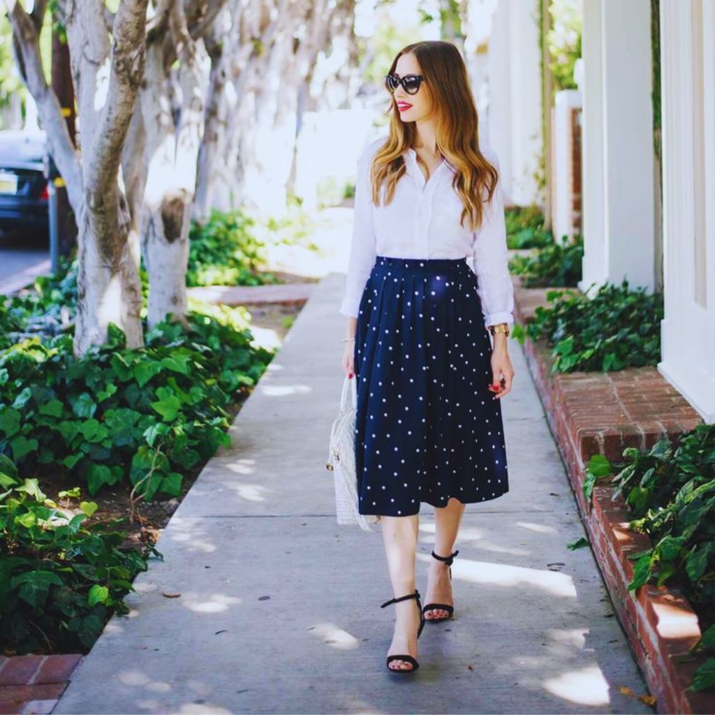 Model in a knee-length polka dot skirt and crisp white shirt, accessorized with minimal gold jewelry — timeless and chic look.
