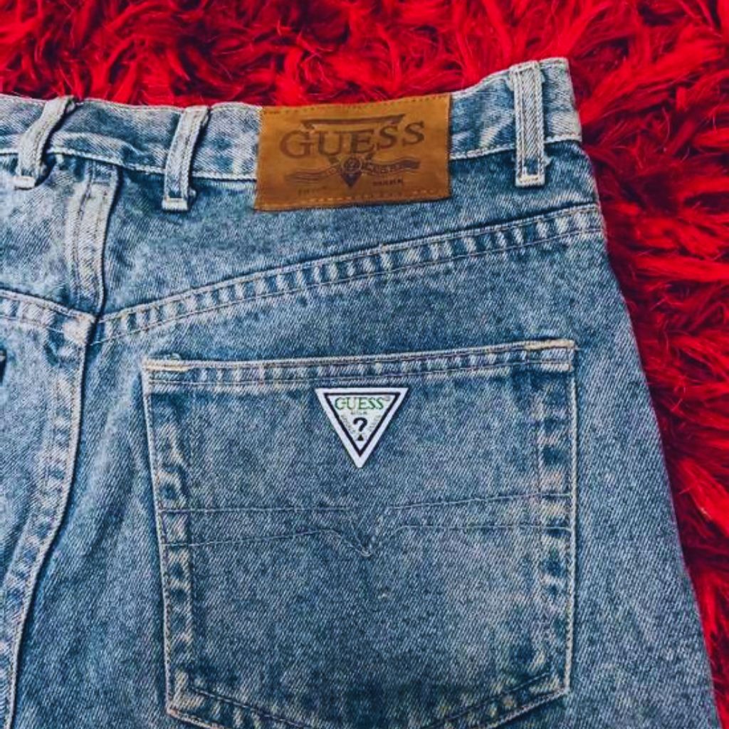 "Vintage Guess jeans with iconic triangle logo on pocket styled with 80s-inspired accessories on flat surface."