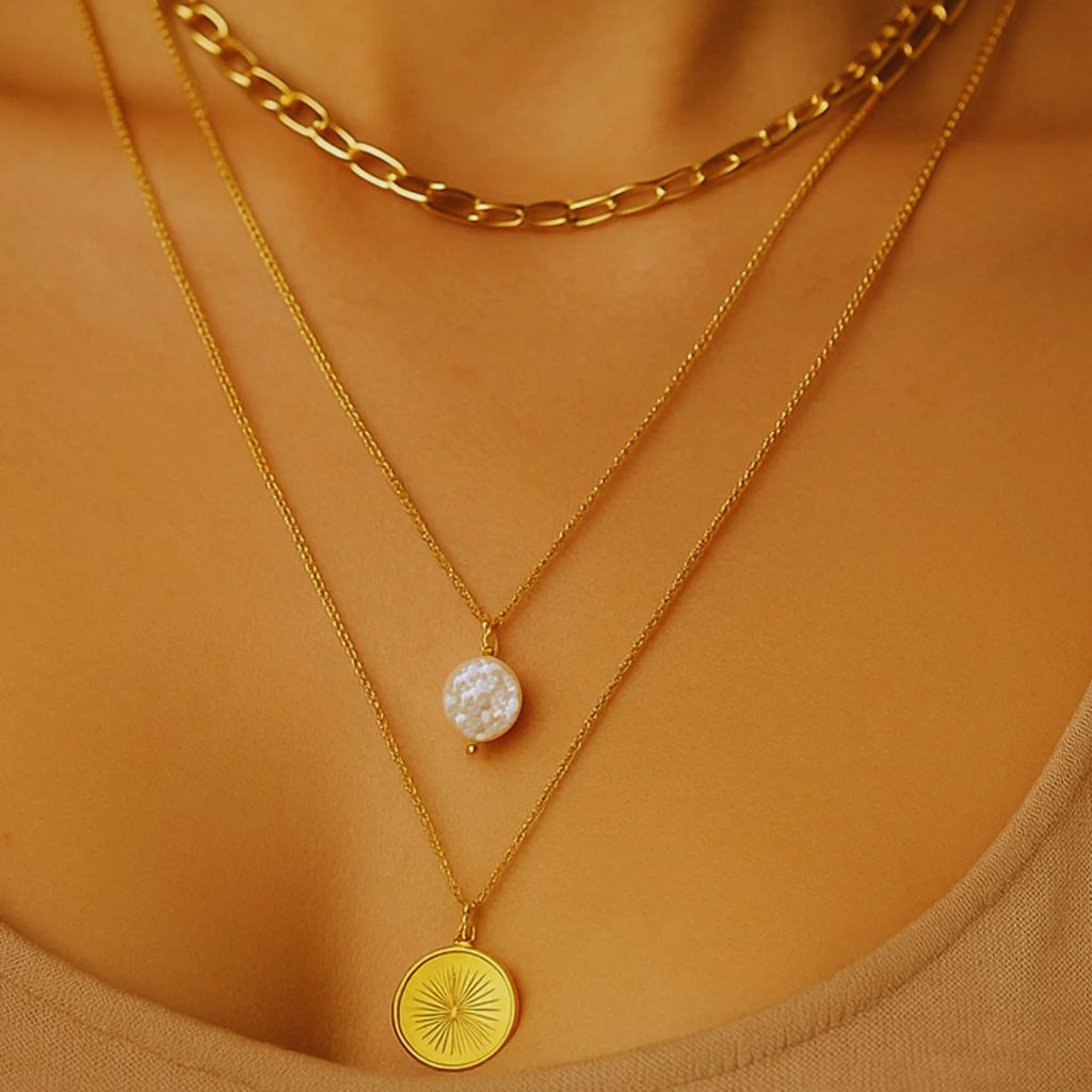 Close-up of a woman wearing layered gold necklaces in varied proportions — thin chain, medium chain, and chunky pendant chain — showing balanced necklace layering.