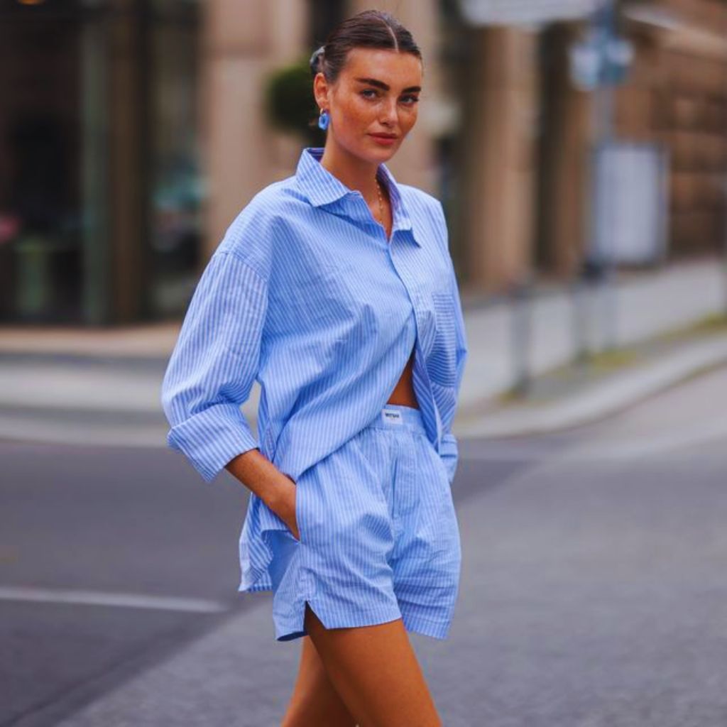 "Woman wearing sky blue shirt and tangerine pants with nude shoes and tote bag standing outdoors in sunlight.