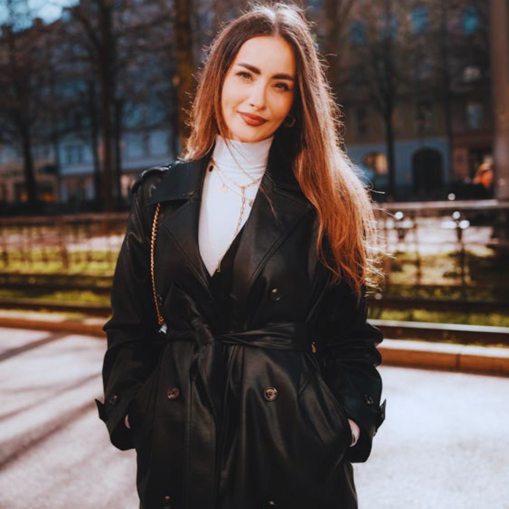 "Woman wearing black leather trench coat layered over hoodie with sneakers posing on city street."