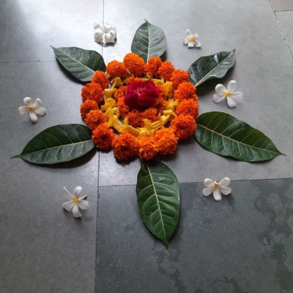A simple flower rangoli design made with marigold flowers, red rose petals, and green leaves, decorated with white plumeria flowers on the floor for Diwali.